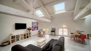 Een ruime woonkamer met een hoge plafond, dakramen, een flatscreen televisie, een boekenkast, een eethoek met een tafel en stoelen, en een bank.