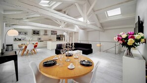 Een ruime loft met hoge plafonds, zichtbare balken, een eethoek met een ronde tafel en stoelen, een keuken op de achtergrond, en een zithoek met een bank en een bloevas.
