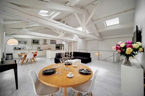 A spacious loft with a high ceiling, exposed beams, a dining area with a round table and chairs, a kitchen in the background, and a living area with a sofa and a vase of flowers. - Immogroom - Luminous spacious T4 apartment - AC (Cannes)