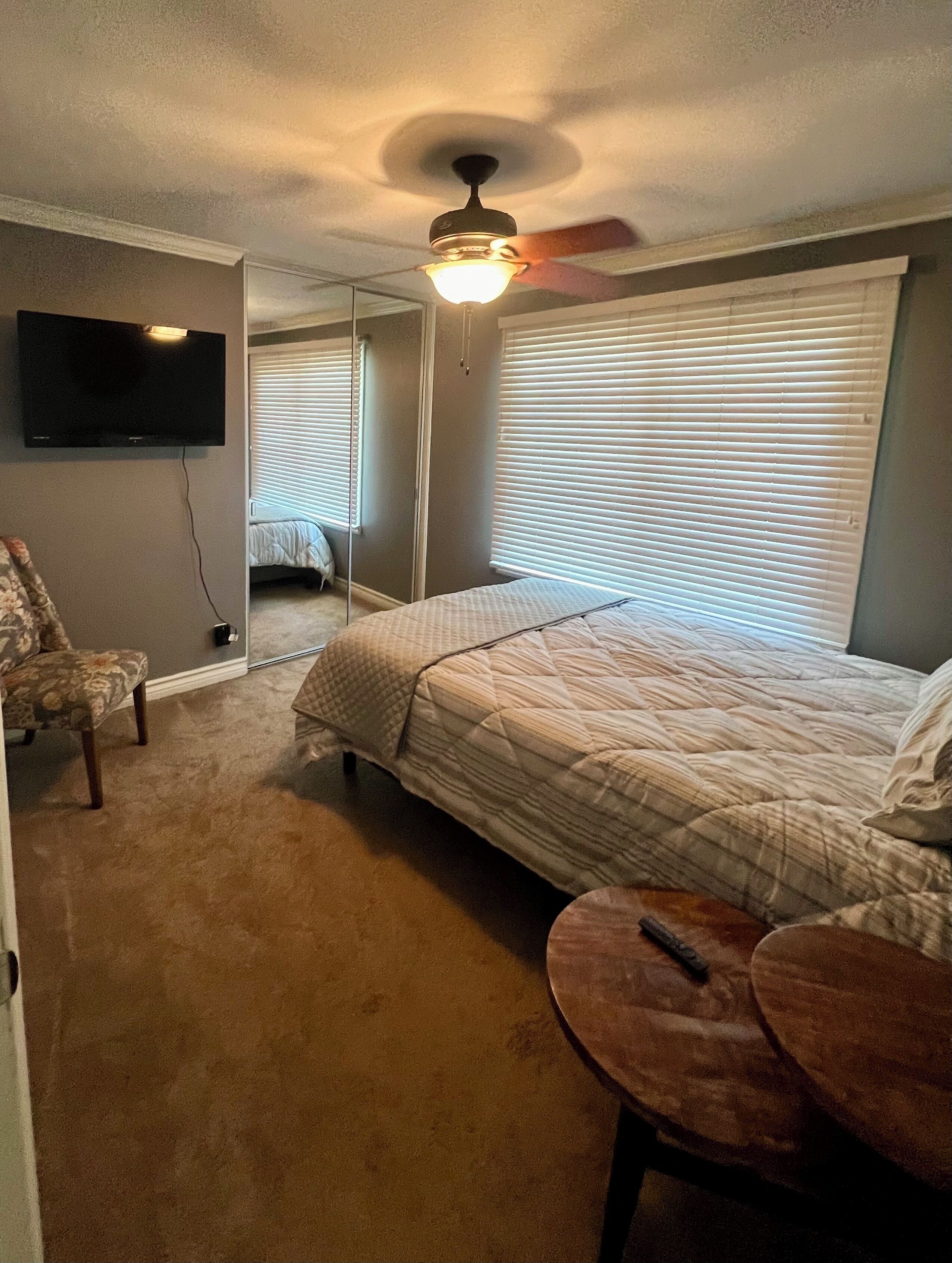 A bedroom with a bed, a chair, a small table, a ceiling fan, a TV, and a window with blinds.