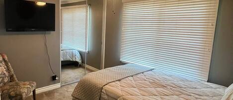 A bedroom with a bed, a chair, a small table, a ceiling fan, a TV, and a window with blinds.