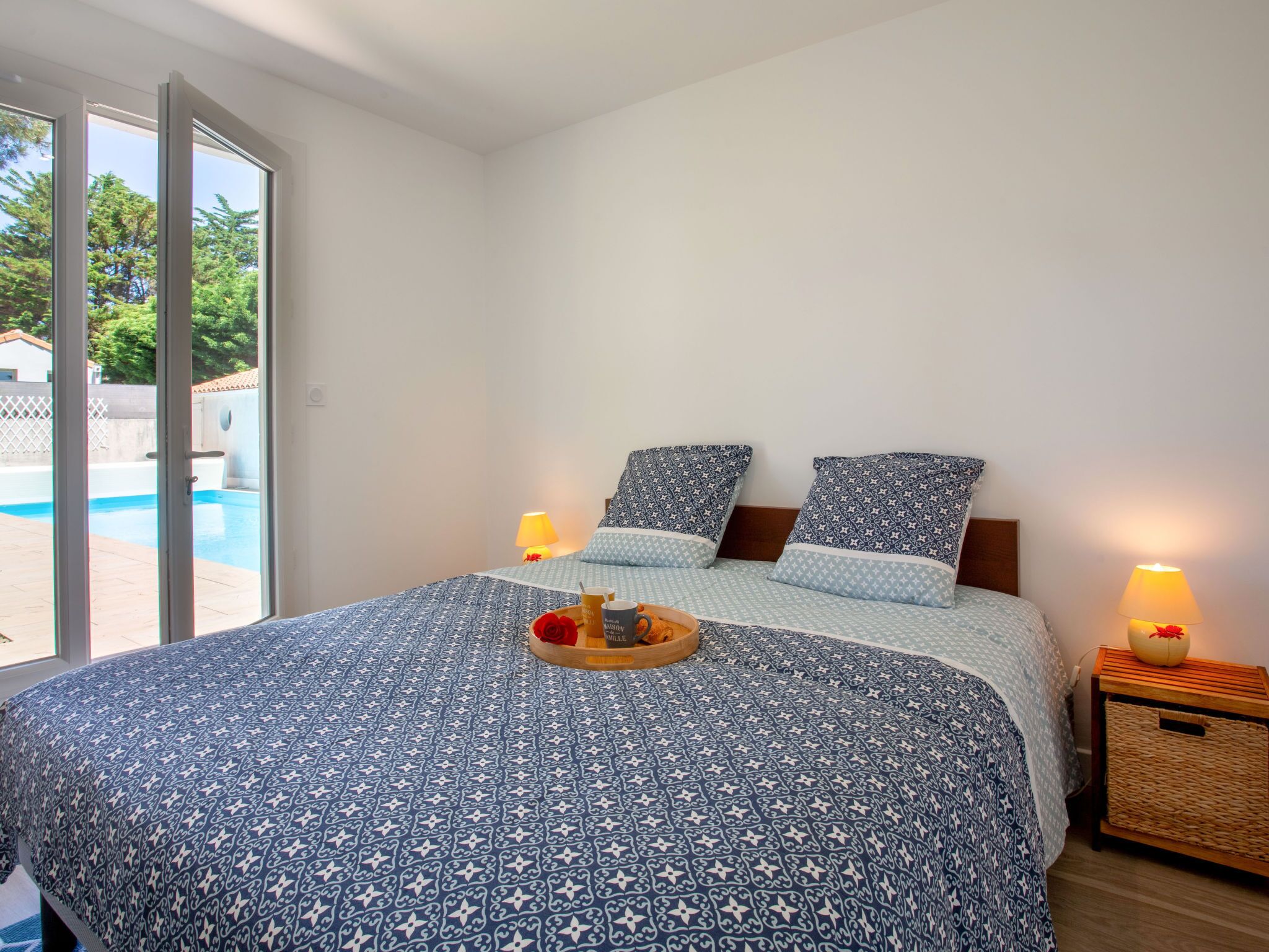 A bedroom with a bed, two bedside lamps, a tray with a drink and a rose, and a view of a swimming pool through a glass door.