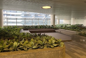 Amplio espacio interior con una jardinera grande llena de diversas plantas, una zona de estar y vistas a edificios residenciales a través de grandes ventanales.