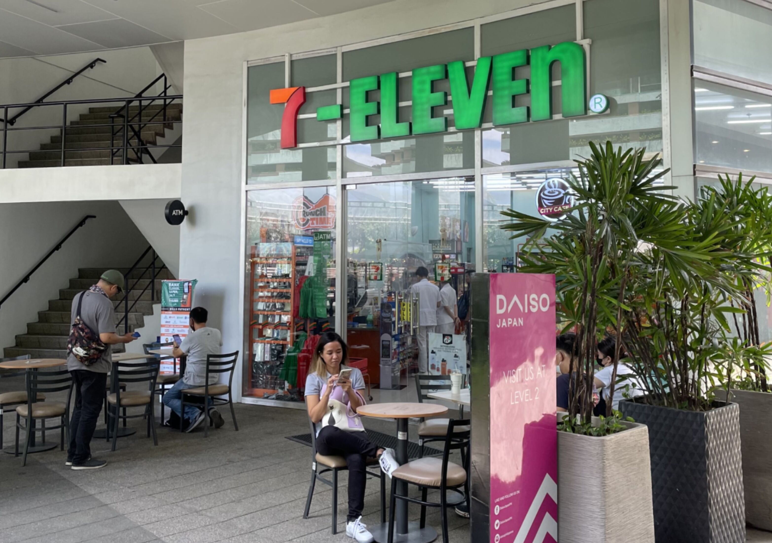 Un magasin 7-Eleven doté d’un distributeur automatique de billets visible, d’un espace avec des tables et des chaises, et d’un escalier menant à un étage supérieur.