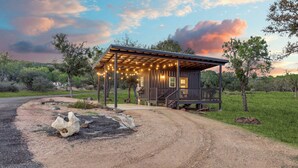 A rustic cabin with a porch, surrounded by a gravel driveway and a grassy area. - The GlampBox. Lakefront, 20acres, Starlink Wifi! (Burnet)
