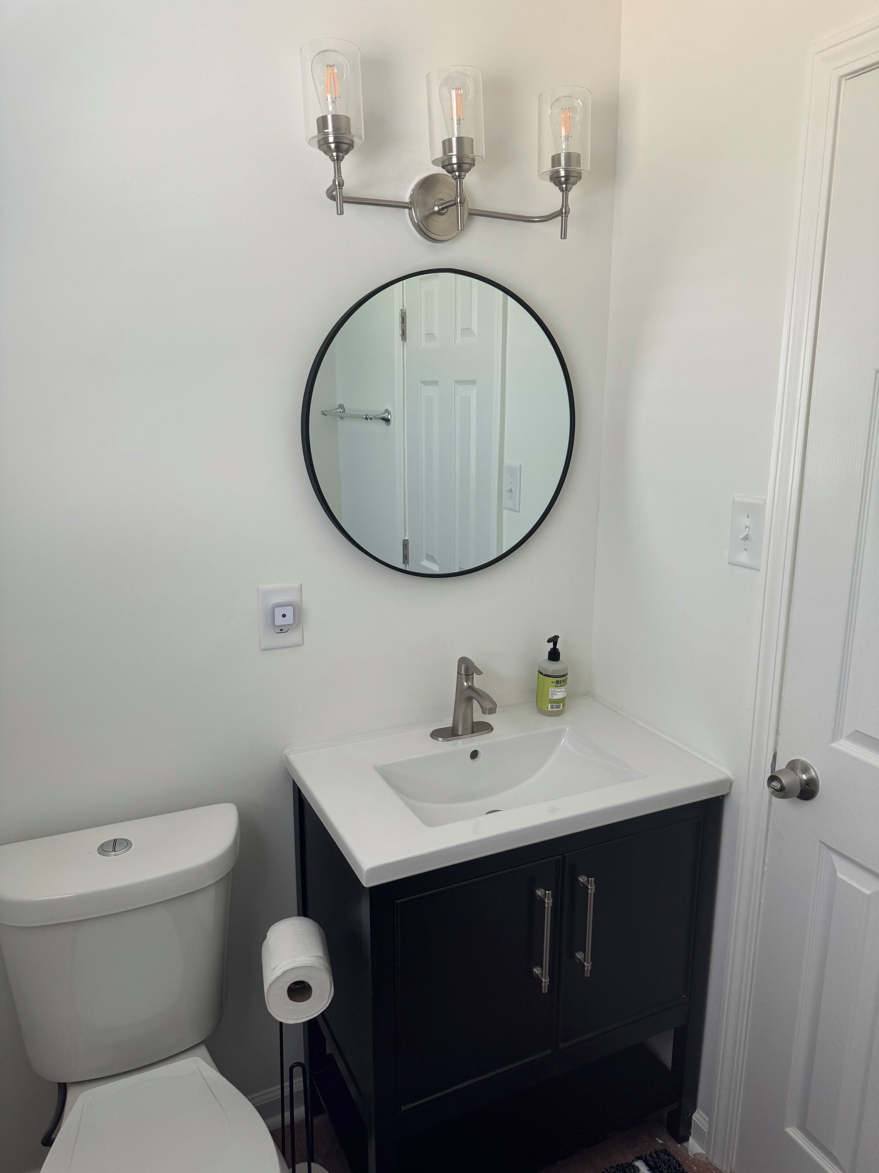 A bathroom with a white toilet, a black cabinet with a sink, a mirror, and a wall-mounted light fixture.