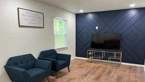 Una sala de estar con dos sillones azul oscuro, un televisor de pantalla plana sobre un pedestal dorado, y una frase motivacional en la pared.