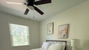 Un dormitorio con cama, ventilador de techo, ventana con persianas, dos cuadros en la pared, mesita de noche con lámpara y alfombra estampada.
