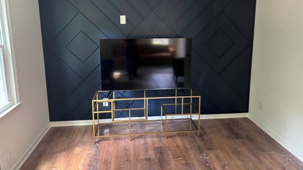 Una habitación con una pared oscura, un televisor montado y un mueble de entretenimiento con marco dorado.