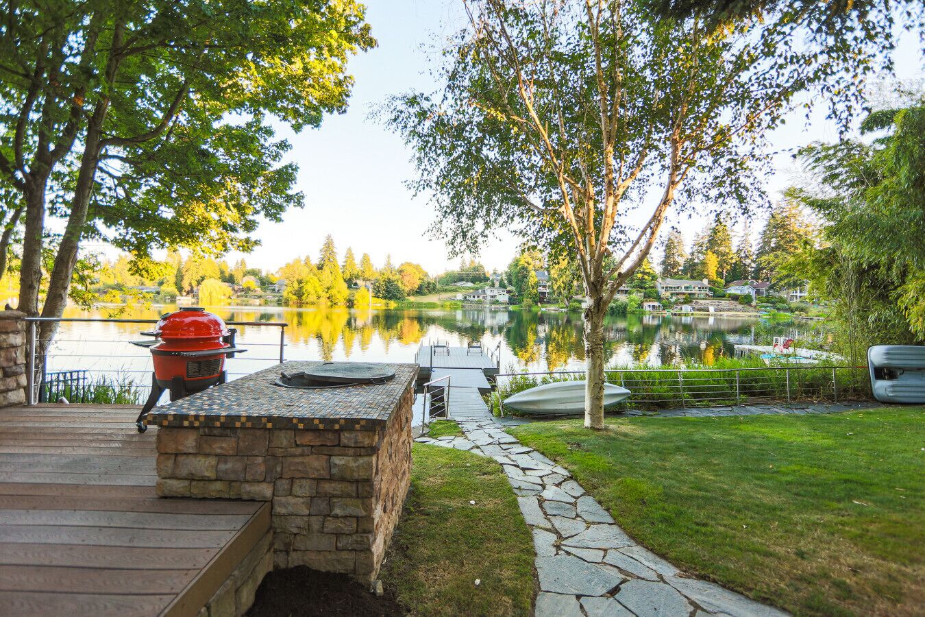 A stone barbecue area with a grill, overlooking a lake with boats and houses in the background.