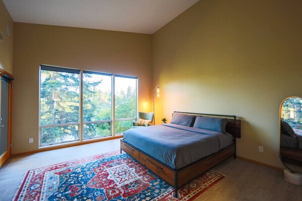 A bedroom with a large window, a bed, a nightstand, and a chair.