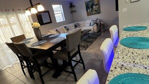 A modern dining area with a white leather couch, a wooden dining table, and a kitchen in the background. - Cozy 3-bedroom townhome inHomestead , near Keys, with AC, full kitchen , WiFi (Homestead)