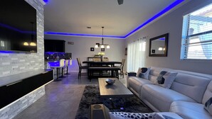 A modern living room with a flat-screen TV, a dining area with a table and chairs, a ceiling fan, and a large window with blinds. - Cozy 3-bedroom townhome inHomestead , near Keys, with AC, full kitchen , WiFi (Homestead)