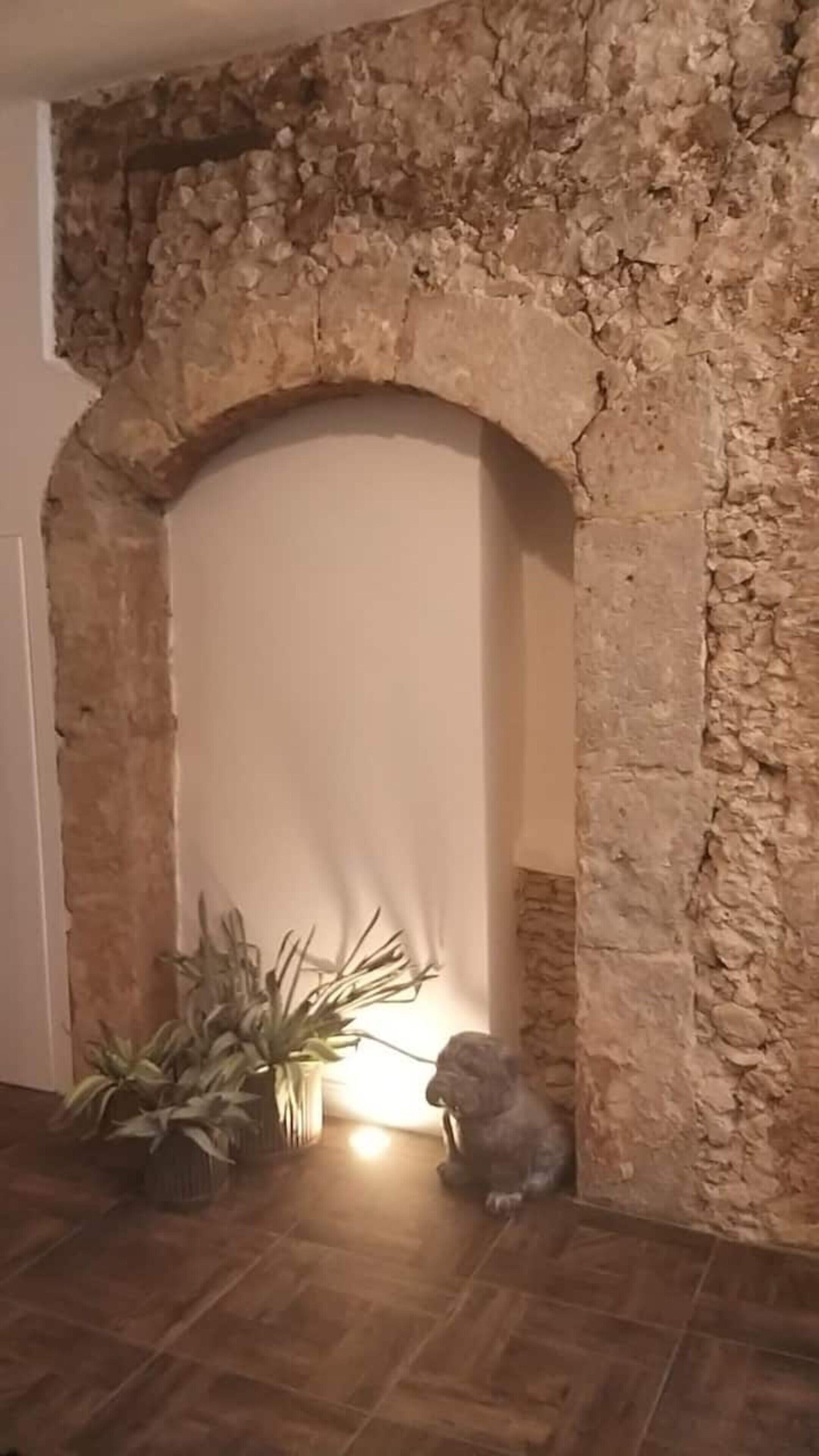 Um arco de pedra que dá acesso a um espaço com soalho ladrilhado, uma planta em vaso e uma estátua.