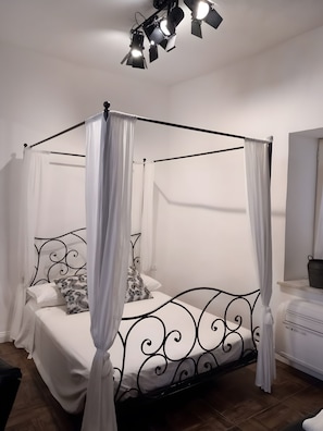 A four-poster canopy bed with white bedding and patterned pillows, a black metal frame, and a ceiling-mounted light fixture.