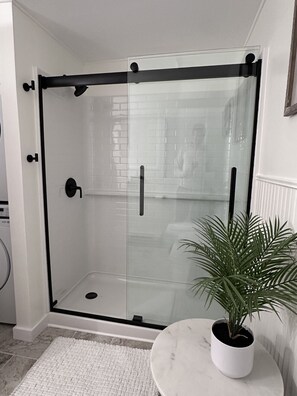 Une douche fermée par des parois de verre, avec un mur recouvert de carreaux blancs, un sol en marbre et une plante en pot posée sur une petite table.