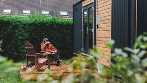 A wooden deck with a table and chairs, a person seated, and a building with a dark exterior and windows. - Garden Home by Tiny Away (Bessé-sur-Braye)
