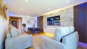 A modern living room with a flat-screen TV mounted on a marble wall, grey upholstered sofas, a dining area with a table and chairs, and a decorative plant. - Romantic flat with Jacuzzi &Pool (Albufeira)