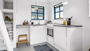 Een compacte keuken met witte kastjes, een houten kruk, een raam met blauwe rand en een klein vloerkleed.