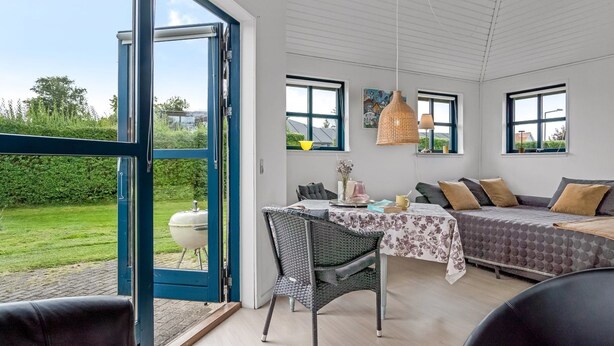 Een moderne kamer met een grote schuifpui naar een tuin, een zithoek met een tafel en stoelen, en een knus zitgedeelte met een bank en kussens.