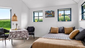 Een slaapkamer met een bed, een tafel en twee stoelen. Er zijn twee ramen met gordijnen, waarvan er een schilderij aan de muur hangt.