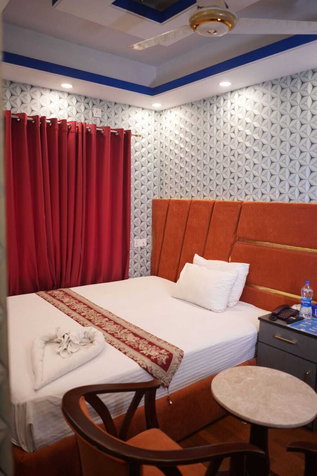 A hotel room with a bed, a nightstand, a chair, a small table, and a red curtain.
