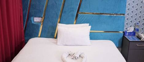 A hotel room with a patterned wall, a bed with a heart-shaped towel, a red curtain, and a blue headboard.