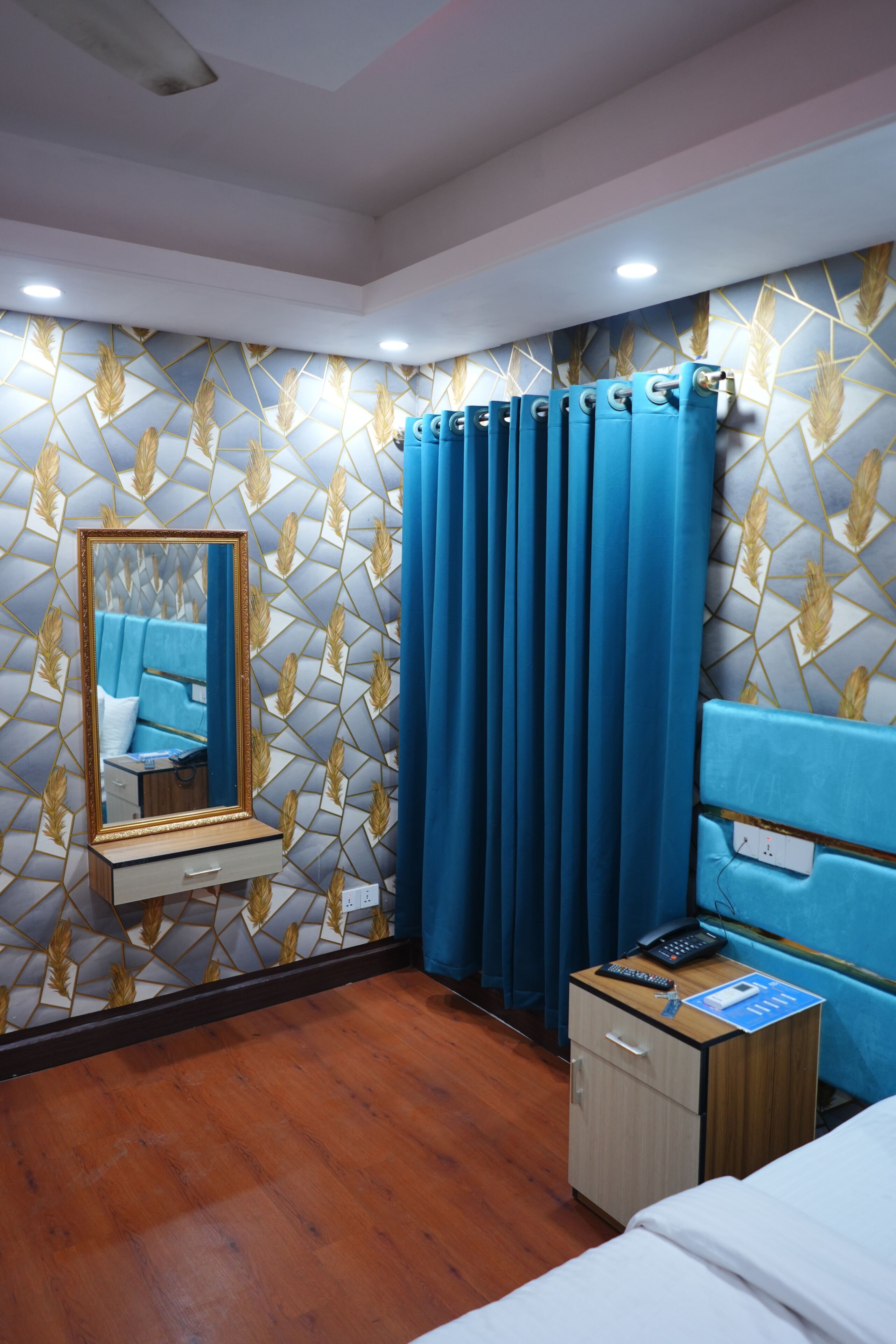 A hotel room with a wooden floor, a bed, a bedside table with a phone and remote, a mirror, and a curtain.
