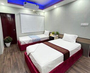 Ein Hotelzimmer mit zwei Betten, einem großen Fenster, einer Deckenklimaanlage, einer Zimmerpflanze und einem Nachttisch mit Lampe.