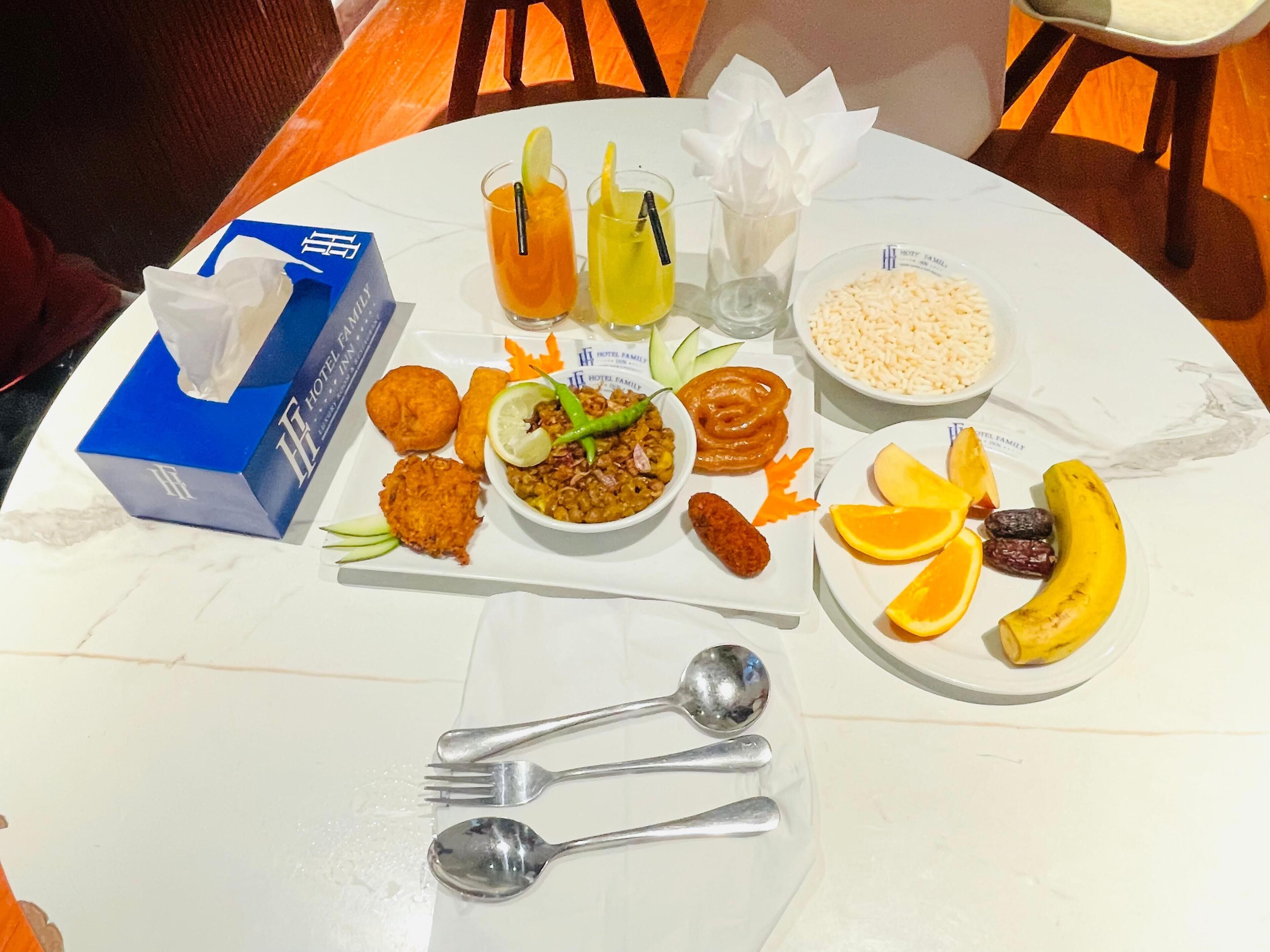A table with various dishes, including rice, curry, and snacks, along with two drinks and a plate of fruit.