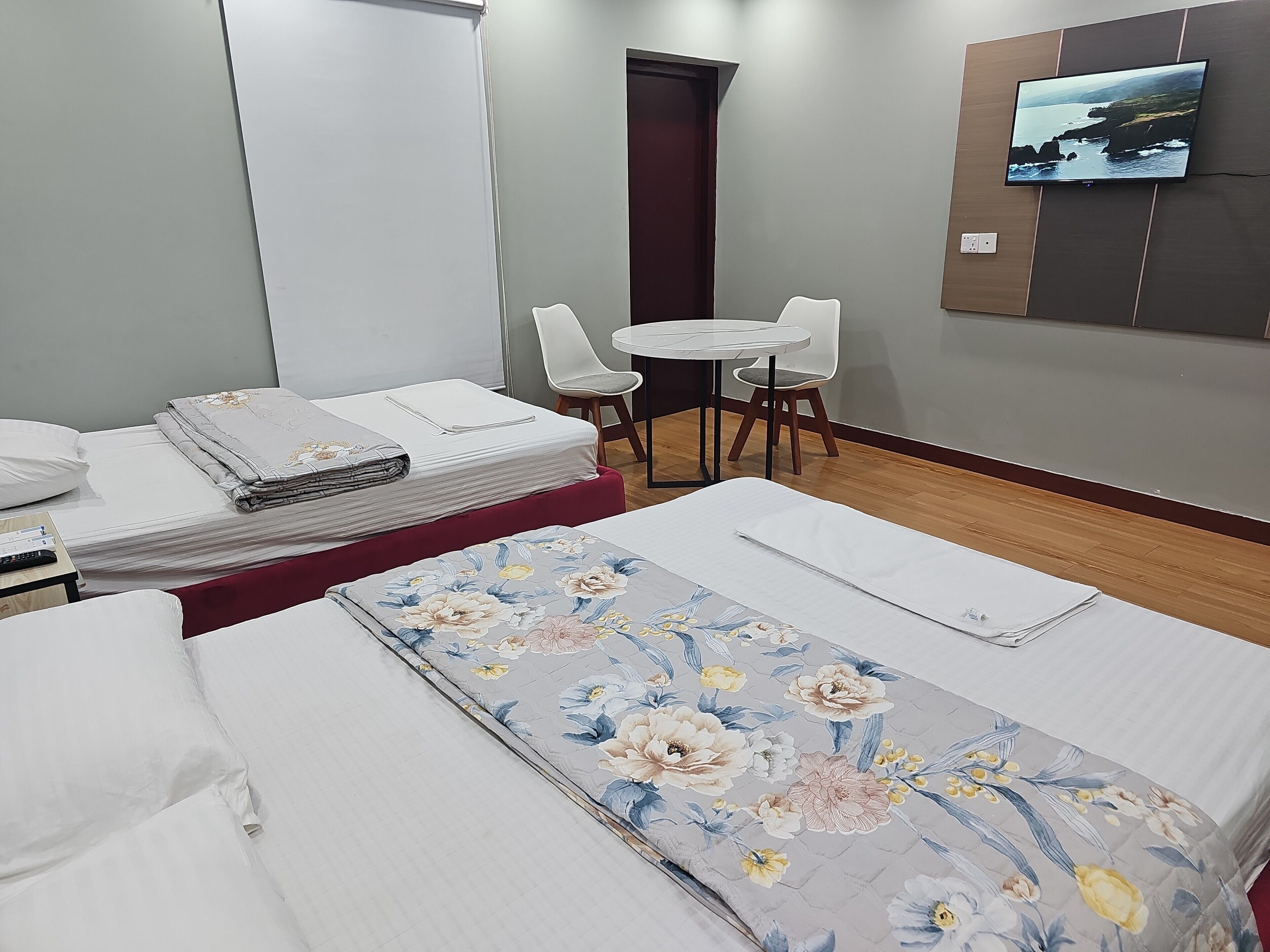 Quarto de hotel com duas camas, uma mesa pequena, duas cadeiras, uma TV fixada na parede e uma porta.