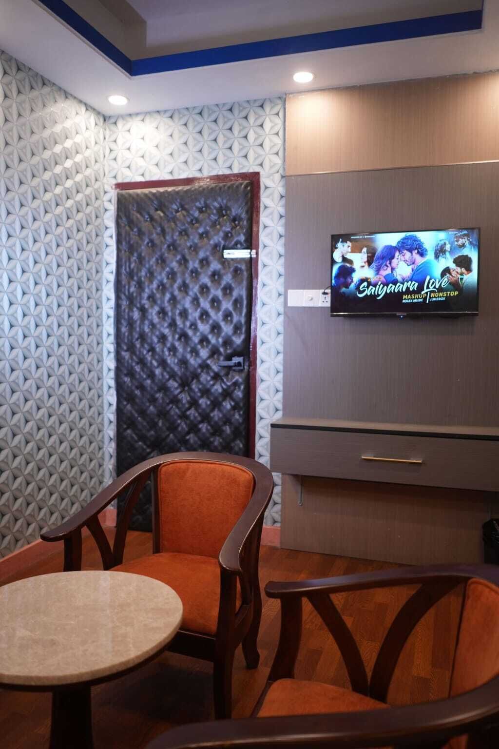 Una habitación con televisión mostrando 'Sanjivana Love', una pared con diseño, una puerta y una zona de estar con mesa y sillas.