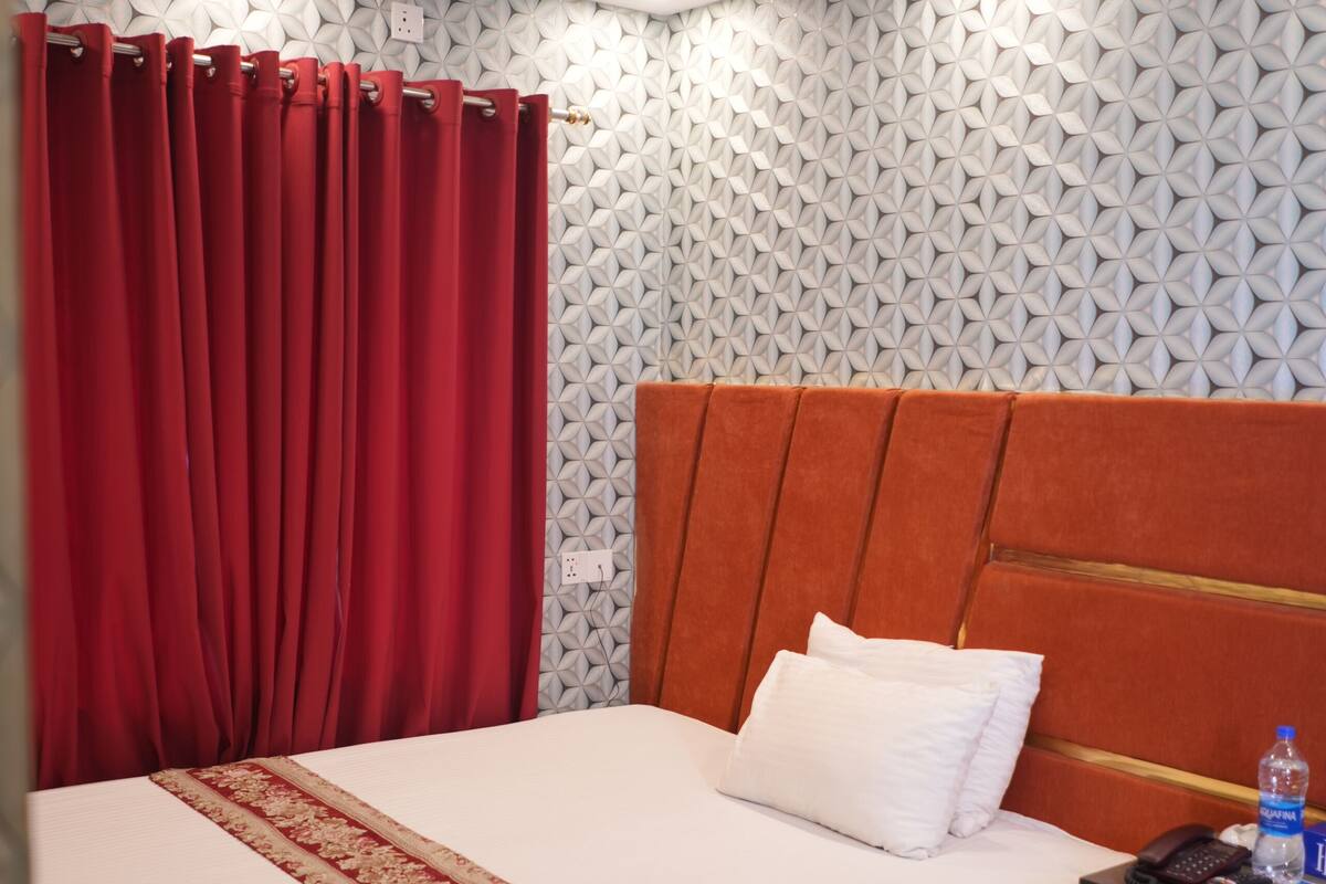 A hotel room with a bed, a small table, a chair, a nightstand, and a red curtain.