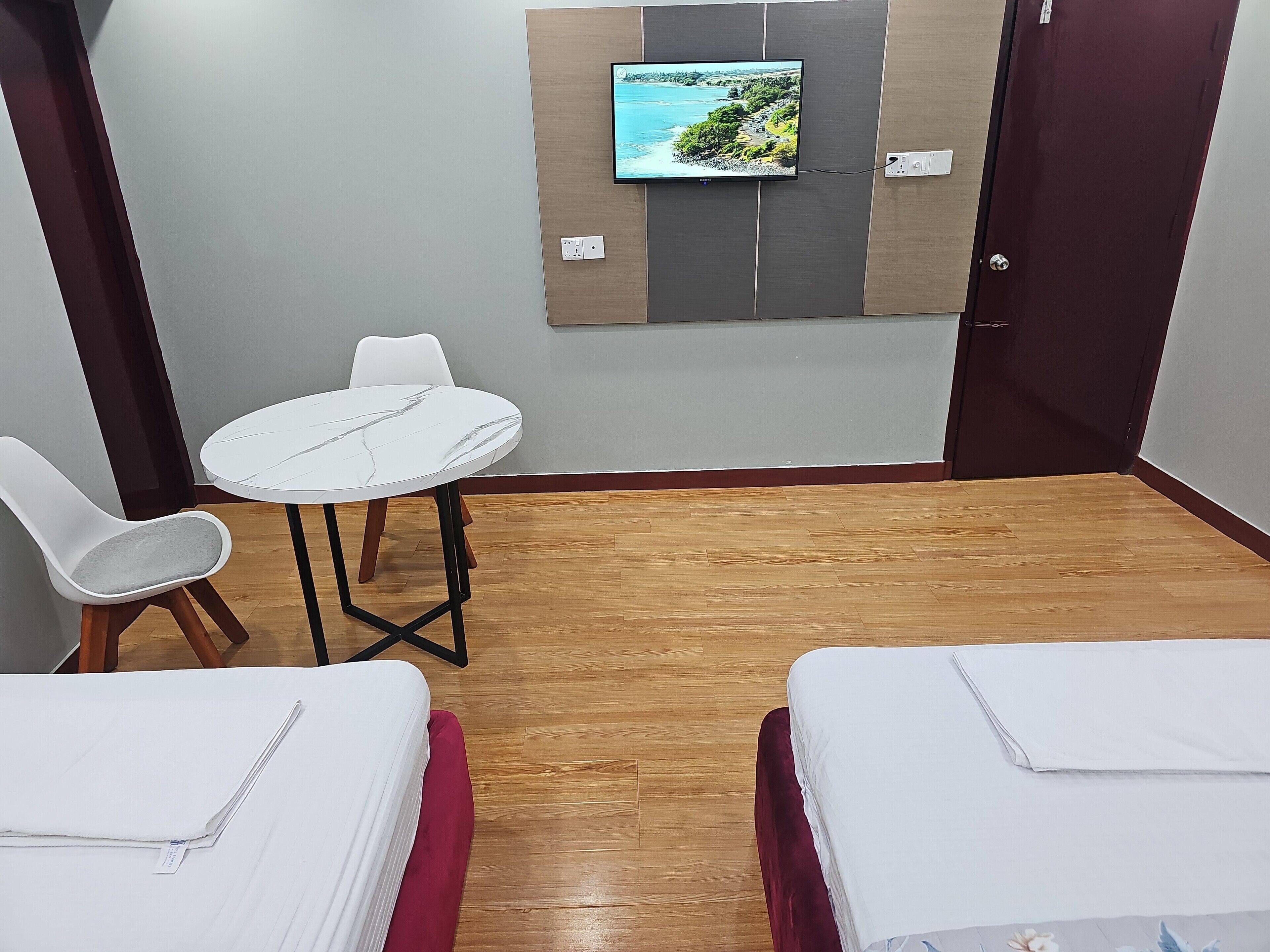 Habitación de hotel con piso de madera, una mesa redonda con una silla blanca, dos camas con ropa de cama blanca y un televisor montado en la pared que muestra una vista panorámica.