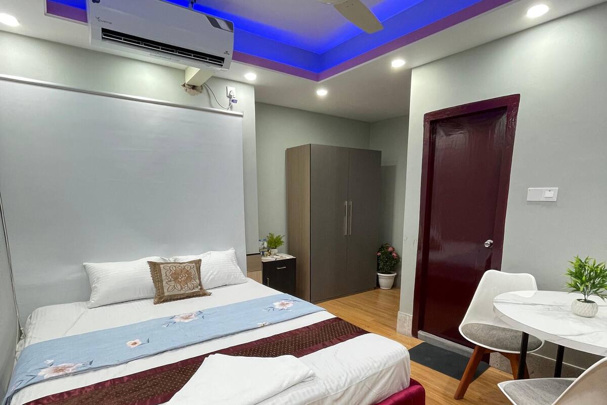 A modern bedroom with a bed, bedside table, air conditioning unit, ceiling fan, and a small dining area with a table and chairs.