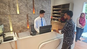 A reception desk with a marble wall, a computer, and a telephone. There is a shelf with various items in the background. - Hotel Family Inn (Dhaka)