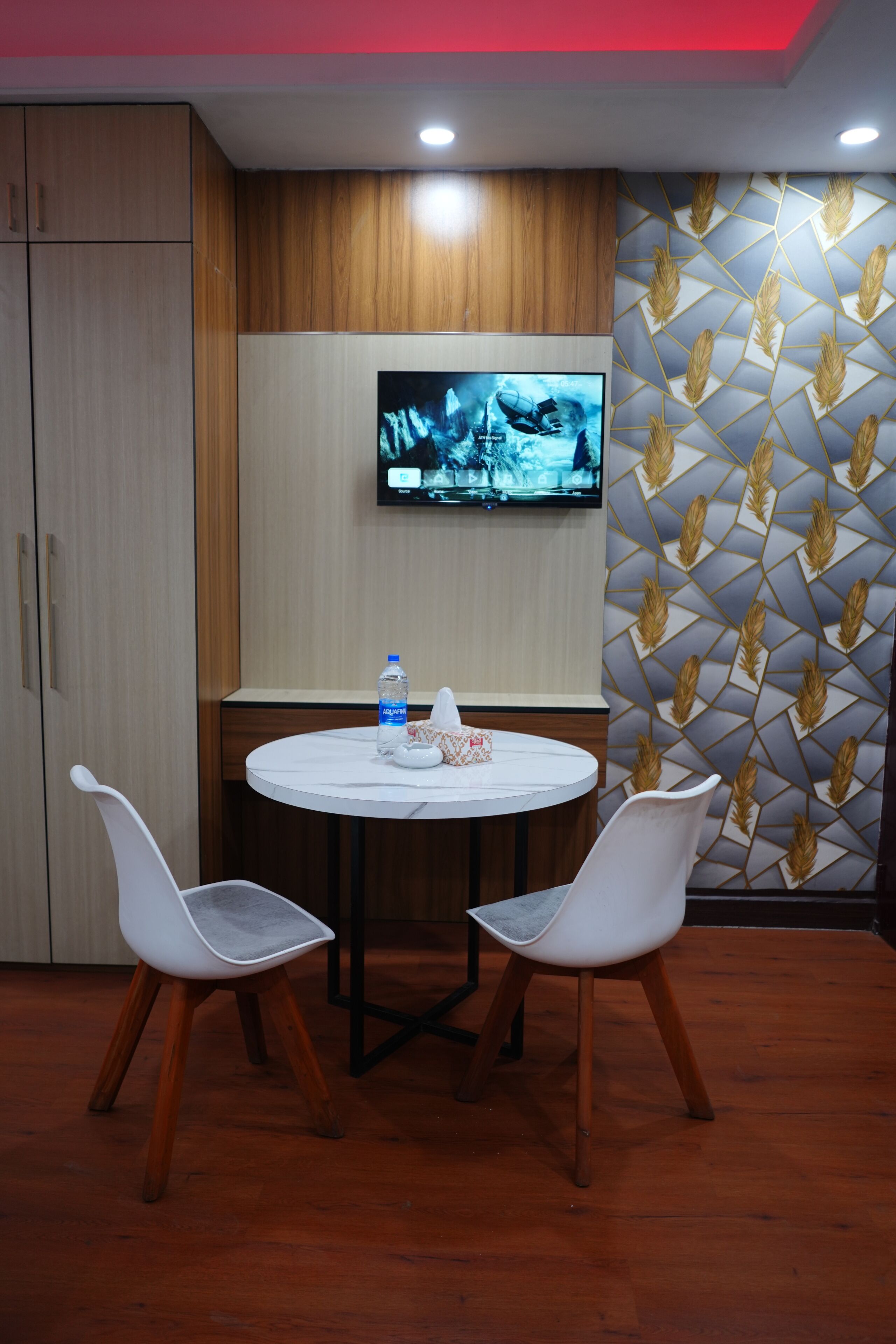 A conference room with a round table, two white chairs, a television mounted on the wall, and a patterned wallpaper design.