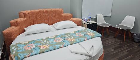 A bed with a floral bedspread, two white pillows, and an orange headboard. There are two white chairs and a desk with a water bottle and a phone.