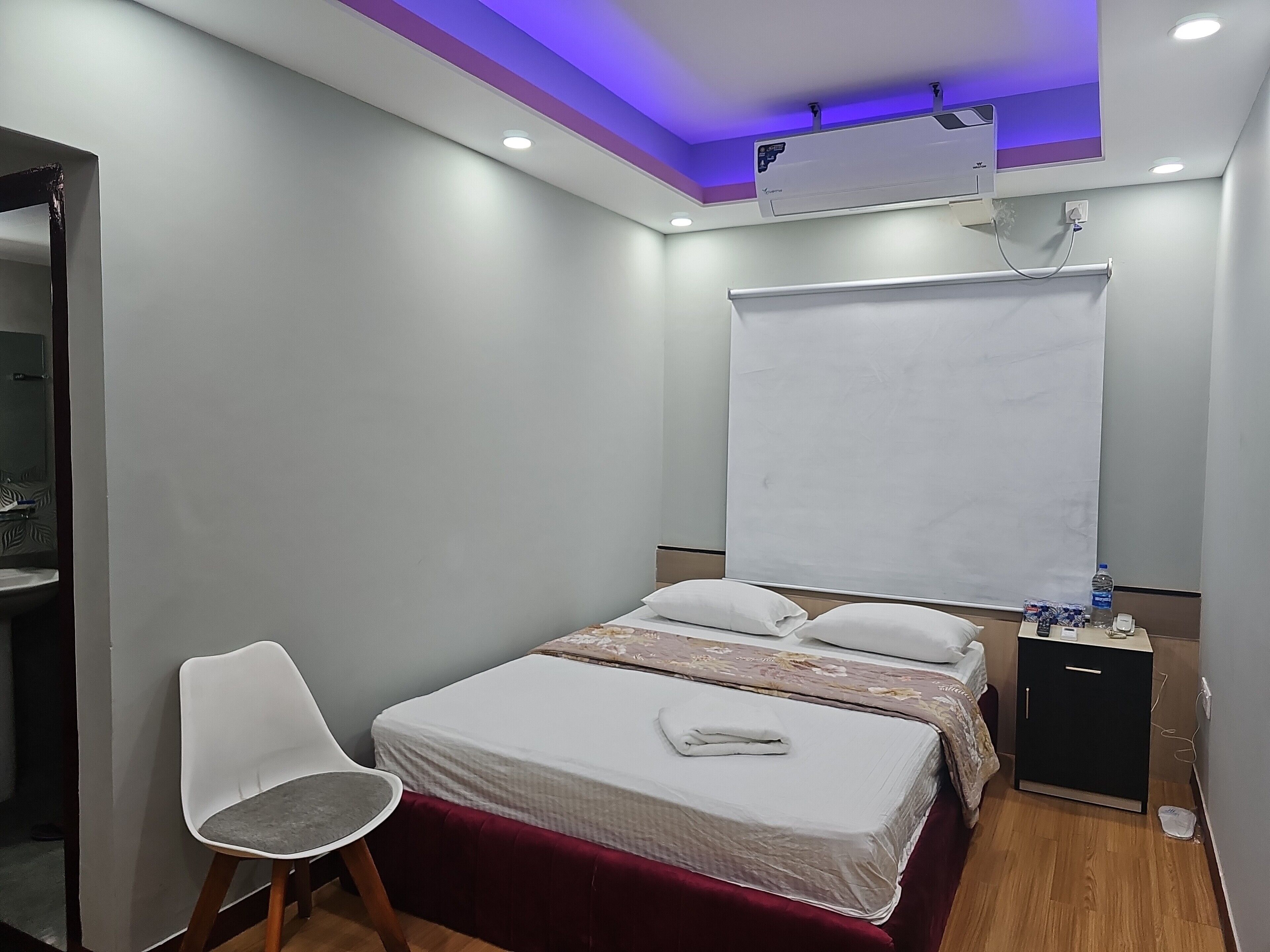 Une petite chambre moderne, équipée d’un lit, d’une chaise, d’une petite armoire et d’un climatiseur fixé au plafond.