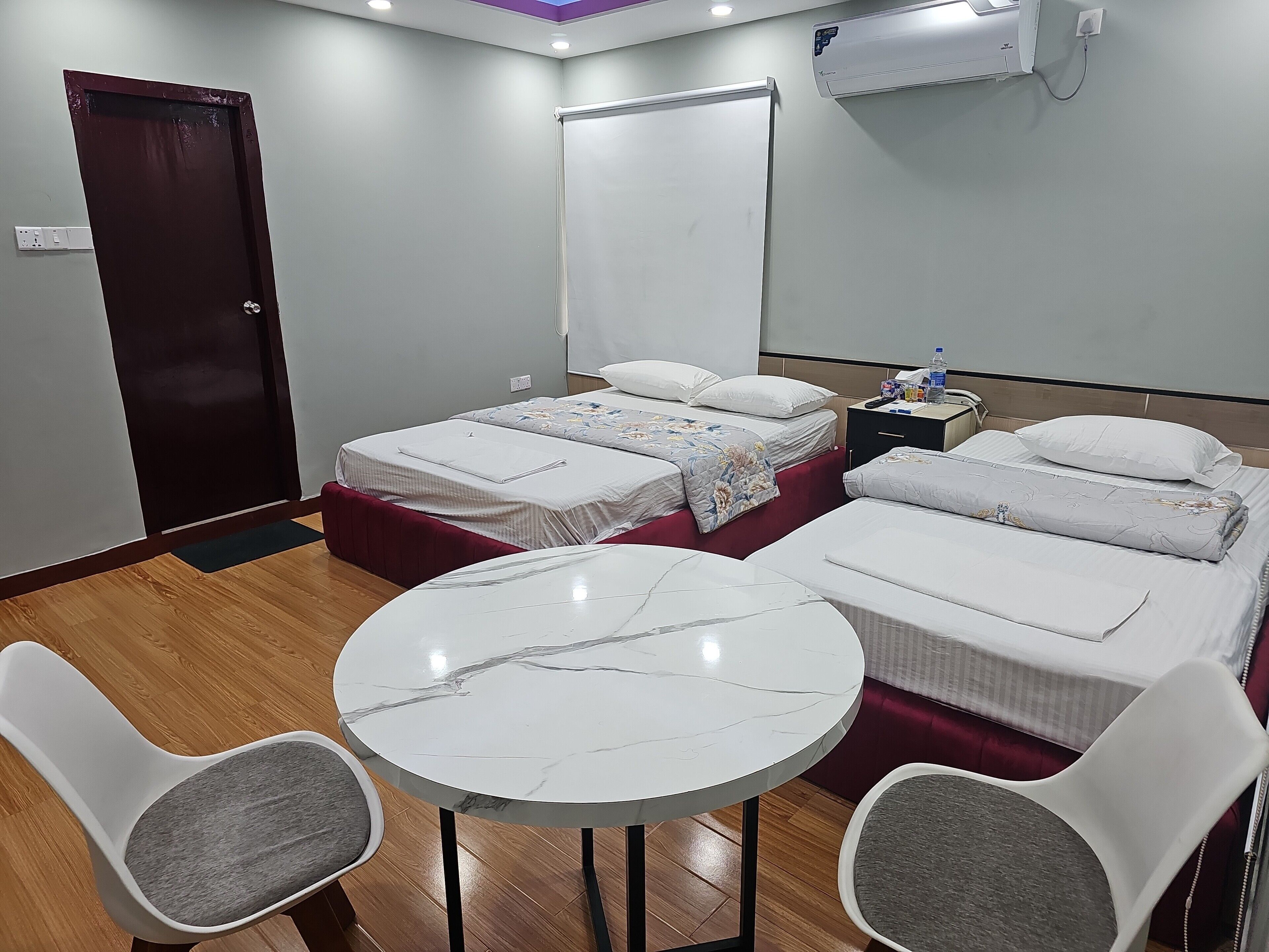 A hotel room with two single beds, a round marble table, two white chairs, a nightstand, and a door.