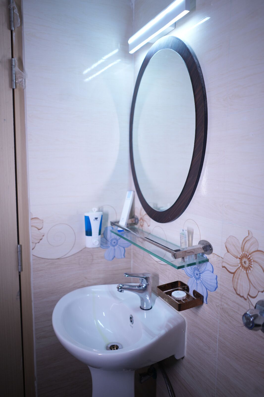 Un baño con lavamanos blanco, un espejo y un estante de vidrio con artículos de tocador.