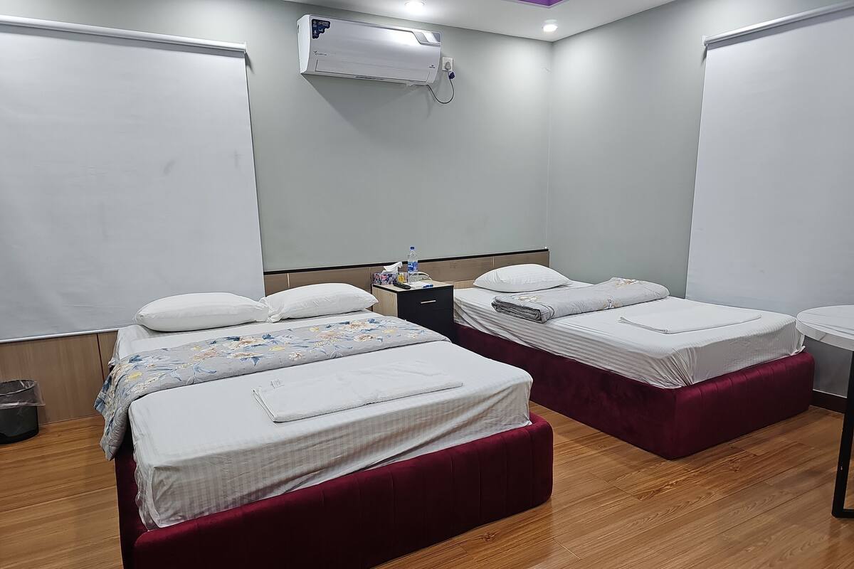 A hotel room with two beds, a wooden floor, a white air conditioning unit, and a small table with toiletries.