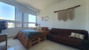 A bedroom with a bed, a wooden nightstand, a bedside table, a brown sofa, and a macrame wall hanging. - Penthouse de lujo (Barranquilla)