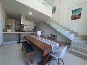 Eine moderne Küche mit einem Holzesstisch, einer Bank und weißen Stühlen. Eine Treppe mit Glastrennwand führt auf eine höhere Ebene.