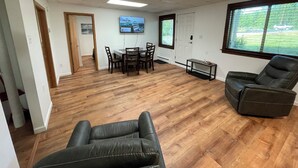 Una habitación amplia con piso de madera, una mesa de comedor con sillas, un sillón de cuero negro, una mesita pequeña y un televisor montado en la pared.