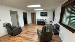 Una habitación con dos sillones reclinables, un televisor montado en la pared, una mesa de comedor con sillas y una ventana con persianas.