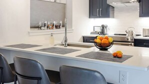 A modern kitchen with a central island, dark cabinets, and a window with blinds. - Stylish 2BR Condo in Newly Restored Church (Charlotte)