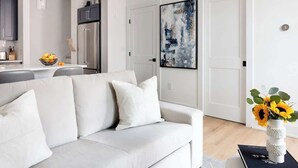 A modern living room with a white sofa, a dining area with a white table, and a kitchen with dark cabinets. - Stylish 2BR Condo in Newly Restored Church (Charlotte)