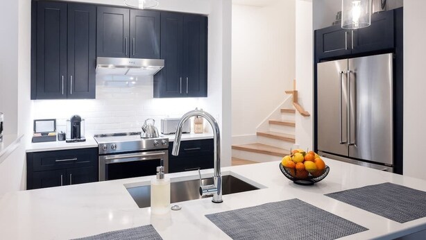 A modern kitchen with dark cabinets, stainless steel appliances, and a central island with a sink and a bowl of fruit. - Stylish 2BR Condo in Newly Restored Church (Charlotte)