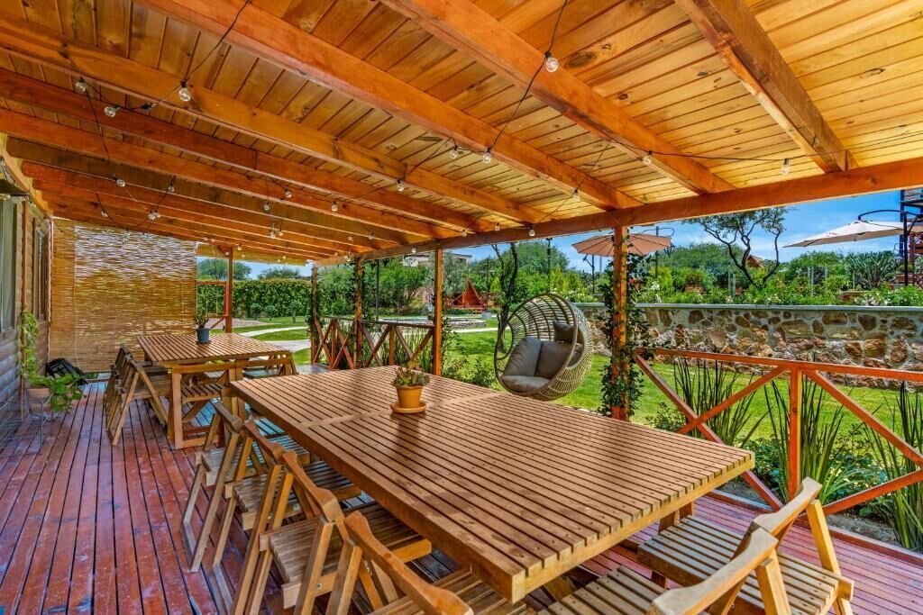 Una terraza exterior de madera con una pérgola, mesas y sillas de madera, una silla colgante y un muro de piedra.